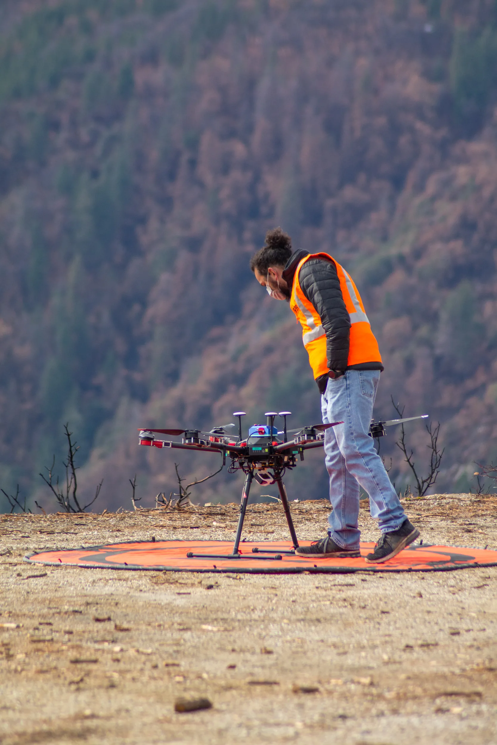 An operator in an orange safety vest looks over a multirotor drone on a landing mat in burned forest terrain.