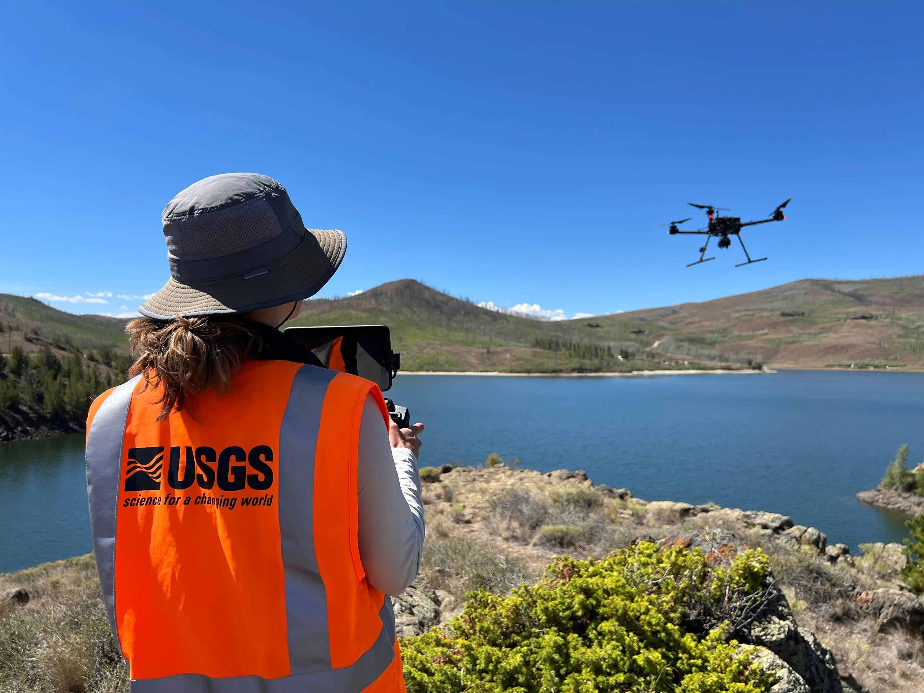 A USGS remote pilot in an orange vest holds a controller while watching a multirotor above a reservoir.