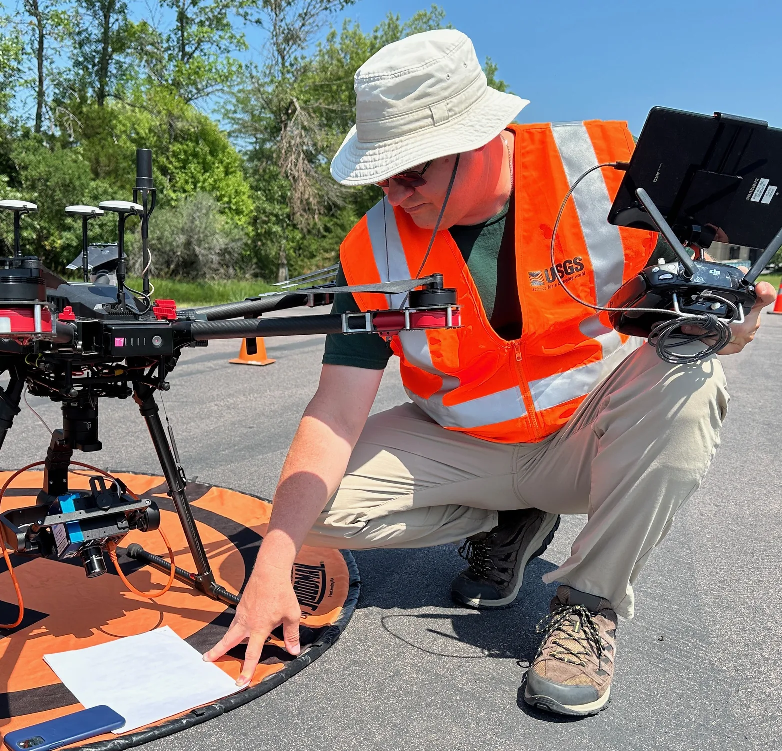 A USGS operator in an orange safety vest kneels beside a drone on a landing pad, holding a controller and reaching toward a paper checklist.