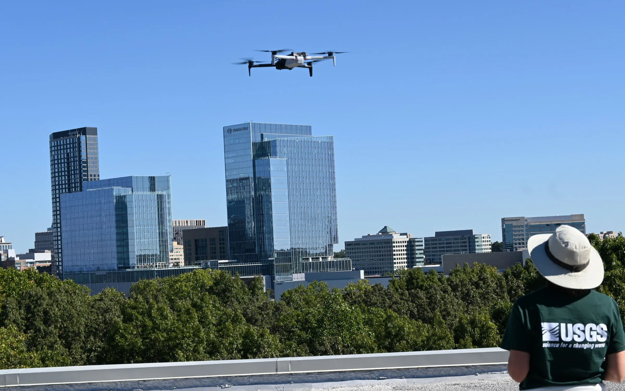 A USGS operator watches a drone flying above a rooftop with city buildings behind it