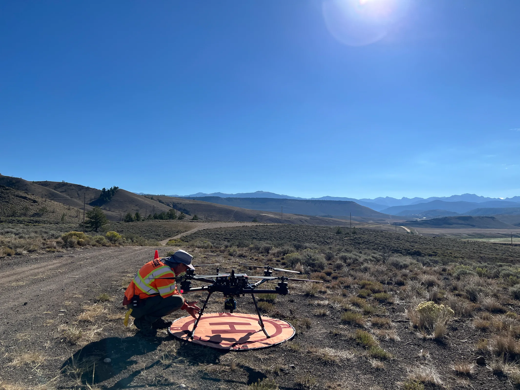 A USGS pilot crouches beside a multirotor drone on an orange landing pad in open terrain near Willow Creek Reservoir.