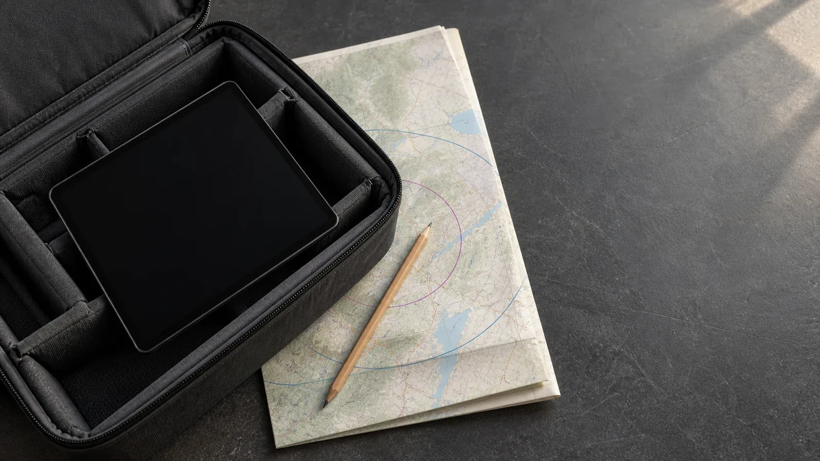 Dark-screen tablet in an open padded case beside an unlabeled planning map and pencil on a charcoal work surface.
