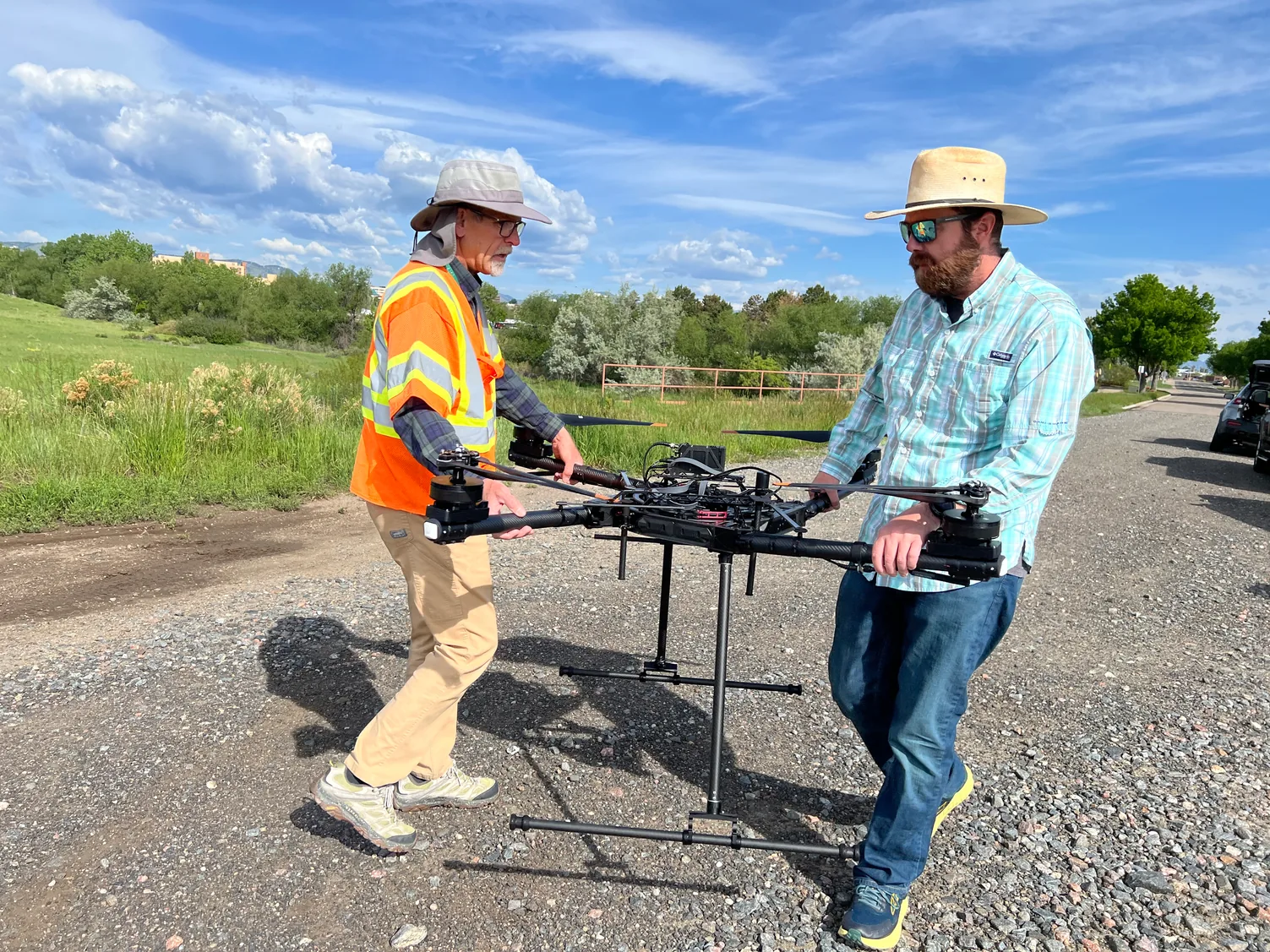 Two USGS remote pilots carry a multirotor drone along a gravel road beside a grassy field