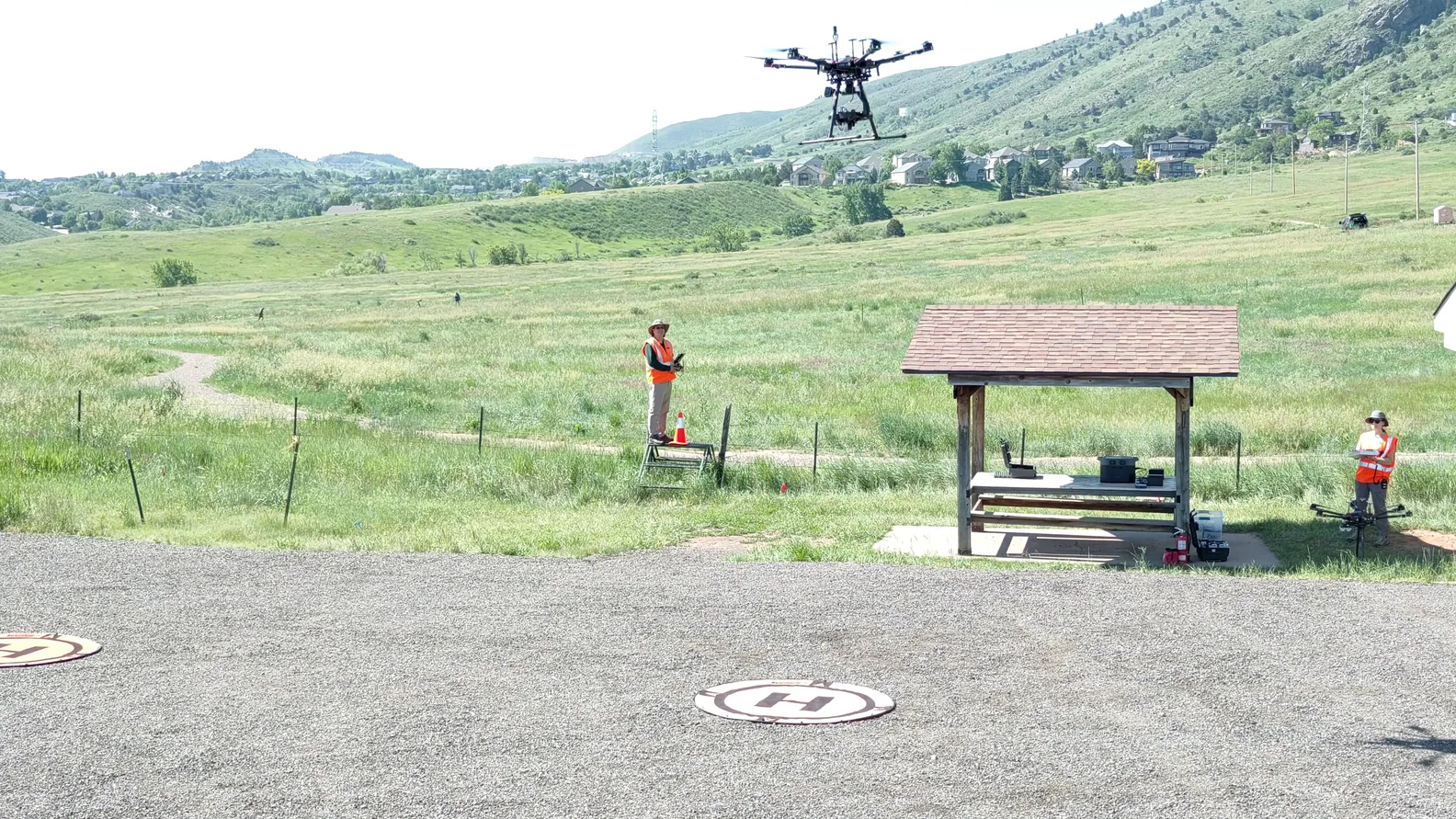 A drone hovers over an open field while two crew members stand near a shelter and landing pads