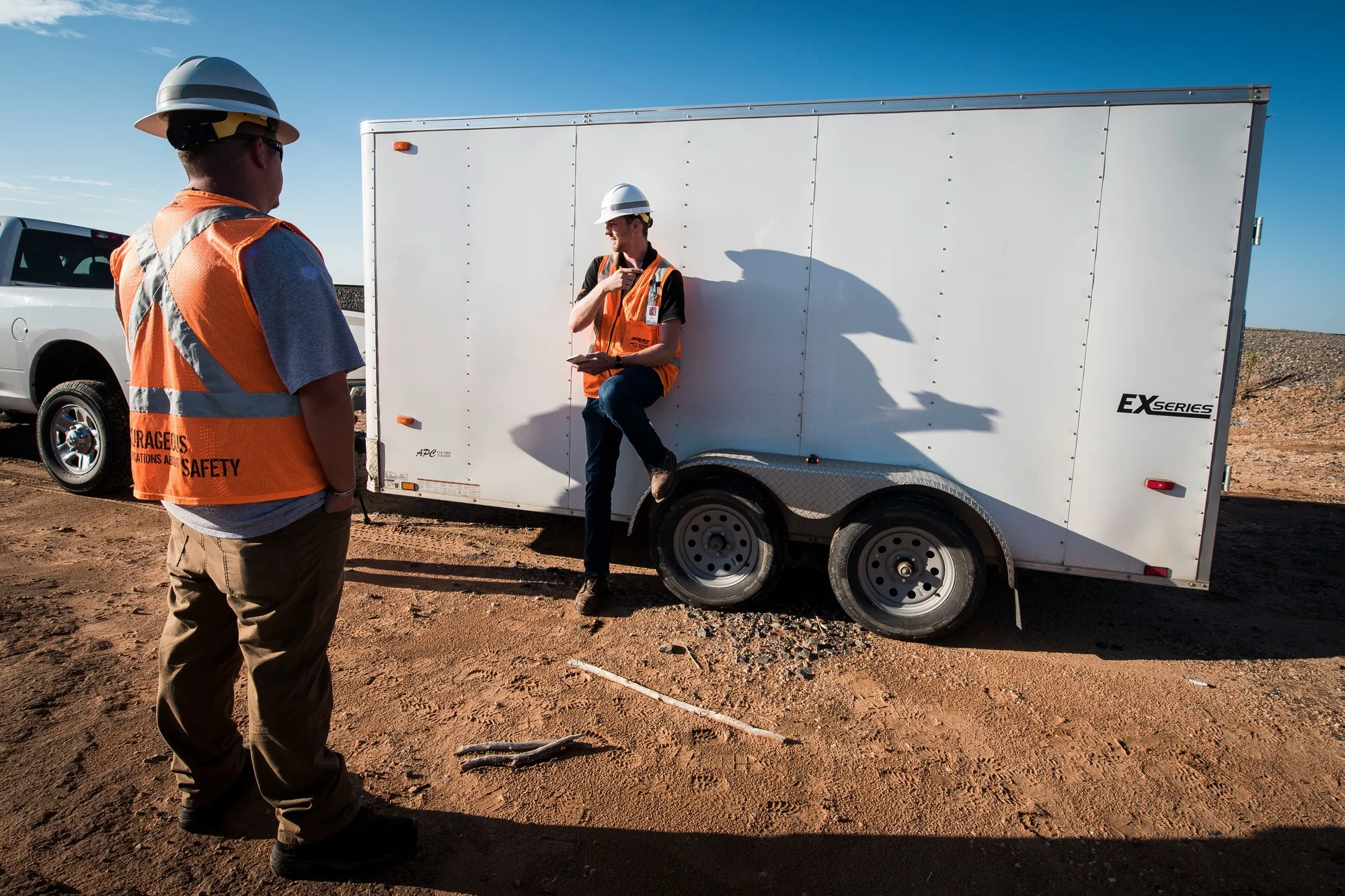 Two drone crew members in hard hats and reflective vests wait beside a field equipment trailer.