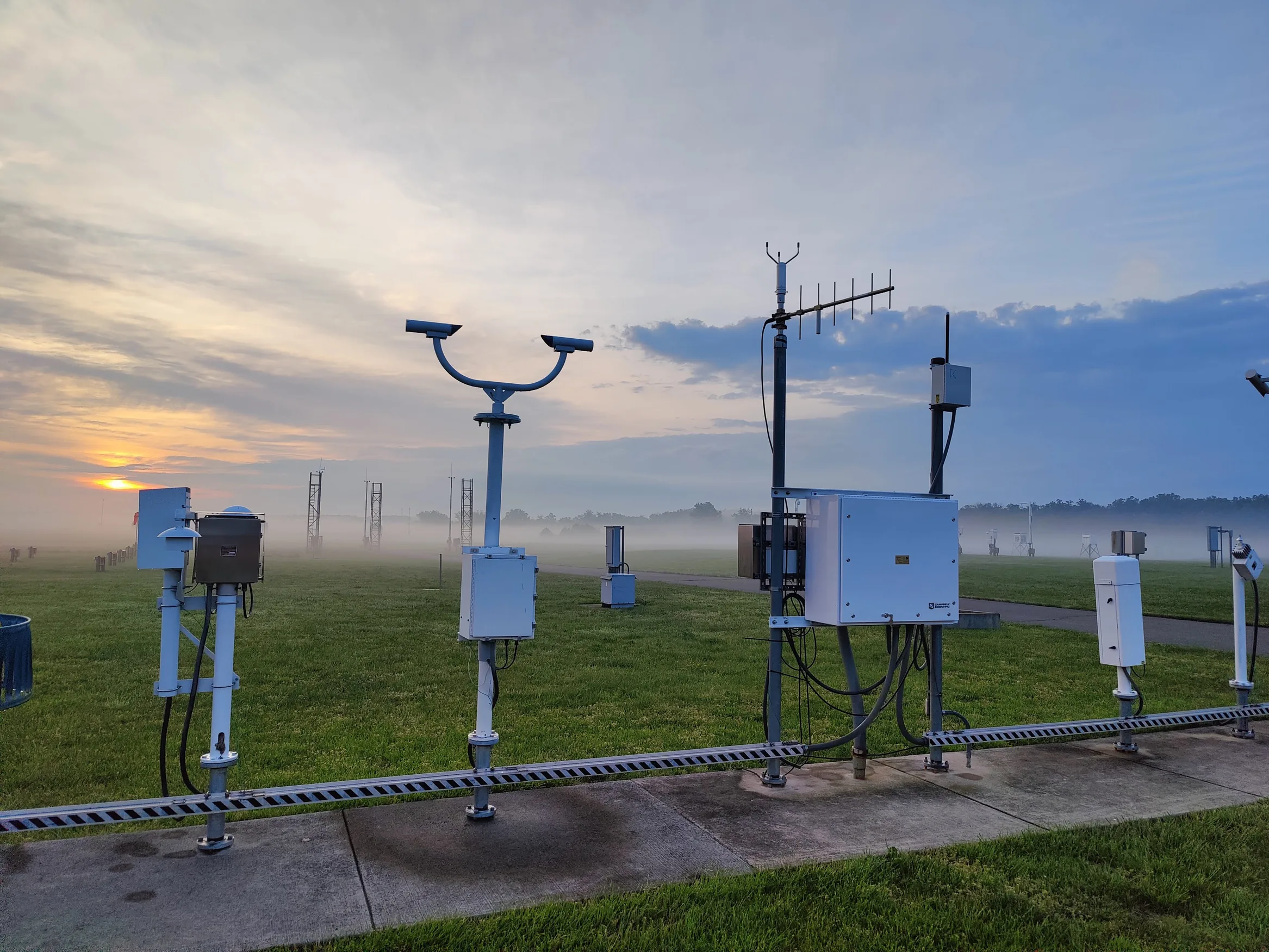 Weather sensors on a grassy observation site, with a low sun and mist in the background.