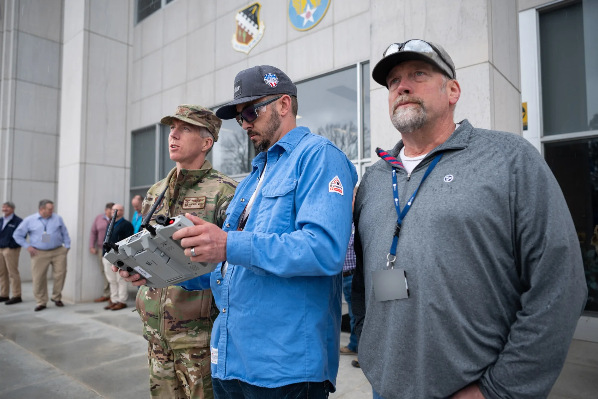 A drone operator holds a controller while two colleagues look toward the aircraft outside a building.
